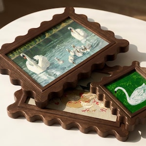 May include: Three framed artworks with scalloped dark brown wooden frames. The top frame features a painting of swans swimming in a lake. The second frame shows a painting of a swan and cygnets. The third frame has a painting of chickens.