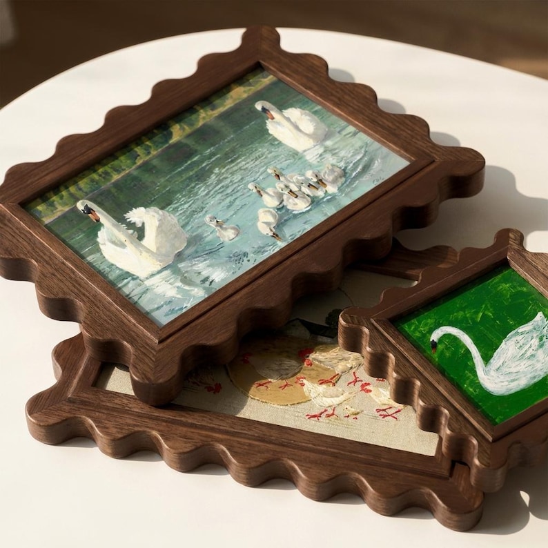May include: Three framed artworks with scalloped dark brown wooden frames. The top frame features a painting of swans swimming in a lake. The second frame shows a painting of a swan and cygnets. The third frame has a painting of chickens.