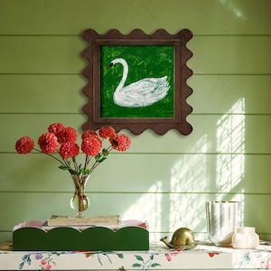 May include: A framed painting of a white swan on a green background. The frame is a dark brown with a scalloped edge. A bouquet of red flowers in a glass vase sits below the painting, along with a green tray and decorative items.
