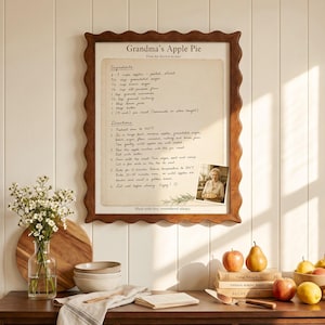 May include: A framed wall art piece titled Grandma's Apple Pie featuring a handwritten recipe with ingredients and directions, accented by a small photograph of a person holding a pie, all encased in a wavy-edged brown wooden frame.