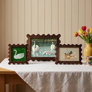 May include: Three framed artworks with scalloped wooden frames. The artworks depict a swan on a green background, swans in a lake, and a farm scene with chickens. The frames are arranged on a white tablecloth.