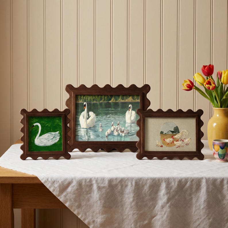 May include: Three framed artworks with scalloped wooden frames. The artworks depict a swan on a green background, swans in a lake, and a farm scene with chickens. The frames are arranged on a white tablecloth.