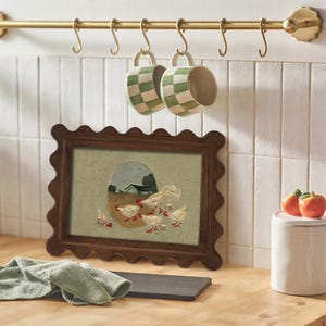 May include: A framed artwork with a scalloped dark brown frame depicts a rooster and chicks in a rural setting. Two green and white checkered mugs hang from a brass bar. A white ceramic jar with a pink lid and two decorative apples are also visible.
