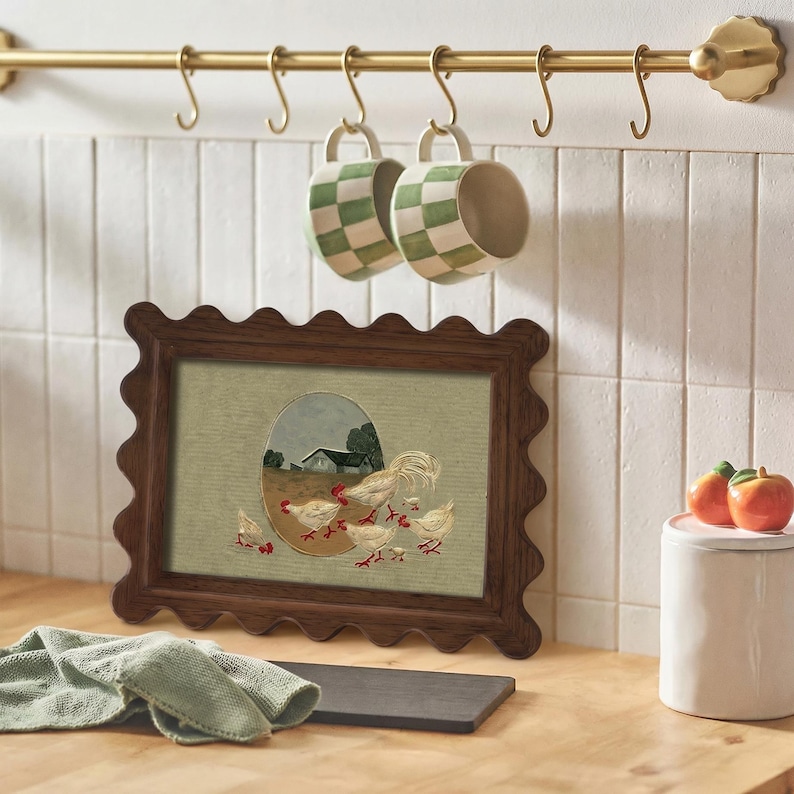 May include: A framed artwork with a scalloped dark brown frame depicts a rooster and chicks in a rural setting. Two green and white checkered mugs hang from a brass bar. A white ceramic jar with a pink lid and two decorative apples are also visible.