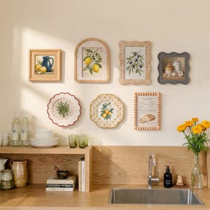 Può includere: Una scena di cucina con quadri incorniciati, piatti decorativi e una mensola in legno. L'arte comprende stampe botaniche di limoni e olive e una natura morta con una brocca blu. I piatti hanno motivi floreali. Un vaso di fiori gialli aggiunge colore.