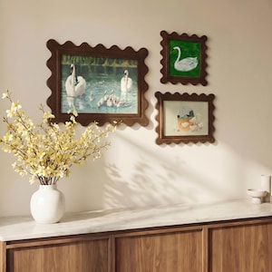 May include: Three framed artworks with scalloped wooden frames are displayed on a wall. The artworks depict swans in a pond, a single swan on a green background, and a swan hatching from an egg. A white vase with yellow flowers sits on a wooden cabinet.