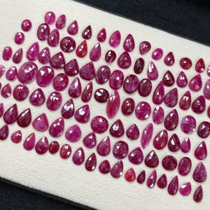 May include: A collection of faceted ruby gemstones in various shapes and sizes, including oval, pear, and round cuts. The rubies display a range of deep red and pink hues, arranged on a white surface.