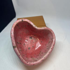 Puede incluir: Un cuenco de cerámica en forma de corazón con un interior rosa y moteado y un borde rosa claro. El cuenco está sobre un bloque de madera. El cuenco es un objeto decorativo.