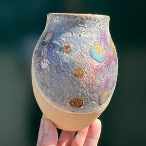 May include: A ceramic vase with a rounded body featuring a textured, iridescent glaze in shades of silver, purple, and blue on the upper portion. The lower section is unglazed, matte tan clay. Small, circular, raised brown and gold accents dot the surface.