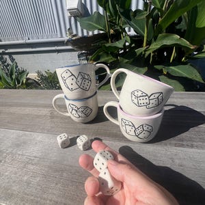 May include: Four white ceramic mugs with black dice illustrations stacked on a wooden surface. Two mugs have blue rims, and two have pink rims. Several white dice with black dots are scattered around the mugs. A hand holds two dice.