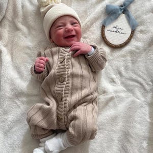 May include: A newborn baby wearing a beige knit romper with wooden buttons and a matching beanie with a pom-pom. The baby is smiling and lying on a soft, white blanket. A circular embroidered name tag with the name "shia woodrow" is also visible.