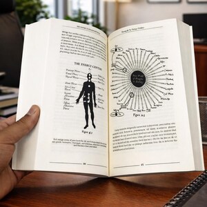 May include: An open book displaying diagrams and text about energy centers, with a human figure illustration and a circular diagram. The book is on a wooden surface, with a hand holding the pages open.