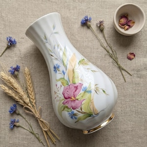 May include: White ceramic vase with a floral design featuring pink and blue flowers, green leaves, and gold accents. The vase is surrounded by dried flowers and wheat stalks, suggesting a rustic or farmhouse aesthetic.
