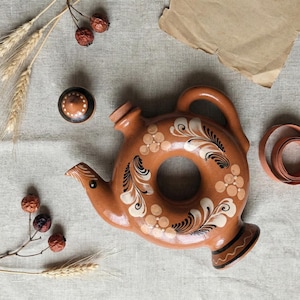 May include: A brown ceramic teapot with a circular body and a handle. The teapot is decorated with white and black floral patterns. A matching lid and other decorative elements are also present.
