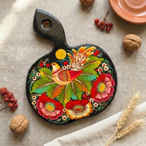 May include: Black wooden cutting board with a handle, featuring a vibrant floral and bird design. The artwork includes red, yellow, and green colors. The board is surrounded by walnuts, berries, and a terracotta plate.