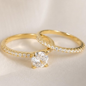 Puede incluir: Un anillo de compromiso de oro con una gran piedra central redonda de talla brillante y una banda pavé, junto a una alianza de oro a juego. Ambos anillos están fabricados en oro amarillo y engastados con numerosas piedras pequeñas, claras y brillantes.