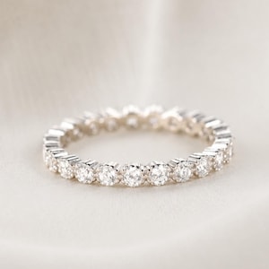 May include: A silver eternity band ring featuring a continuous row of small, round, clear faceted stones set in a metallic frame. The ring has a uniform design with each stone held by small prongs, creating a sparkling, circular band of light.