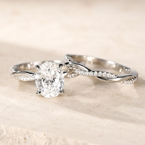 May include: A silver-toned engagement ring set featuring an oval-cut center stone held by four prongs. The band displays a twisted design accented with small, clear pavé-set stones along the shank, paired with a matching twisted wedding band.