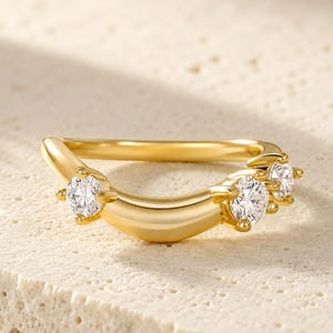 May include: A gold curved ring featuring three round clear stones set in prongs along the front arc. The band has a smooth, polished finish and a distinct crescent shape, showcasing a bright metallic lustre and sparkling faceted gemstones.