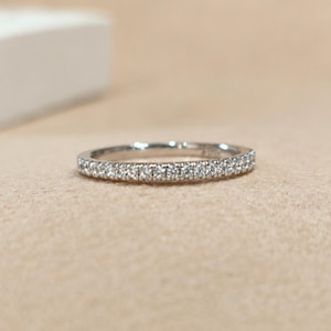 May include: A thin silver-toned band ring featuring a continuous row of small, clear, round-cut stones set along the top half of the metal circumference. The ring has a polished metallic finish and a slim, uniform profile throughout its design.