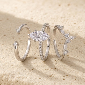 May include: A set of three silver-toned rings. The left ring is an open band with two small round stones. The center ring features a large marquise-cut stone on a pavé band. The right ring is a V-shaped band adorned with multiple small stones.