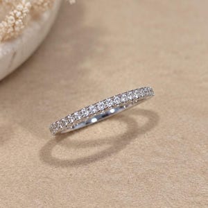May include: A thin silver-toned band ring featuring a continuous row of small, clear, round-cut stones set along the top half of the metal surface. The ring has a polished finish and reflects light across the faceted surfaces of the embedded stones.