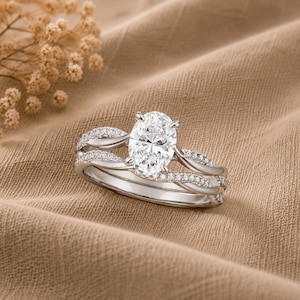 May include: A silver-toned ring set featuring an oval-cut center stone held by four prongs. The band consists of two intertwined strands, with one strand encrusted with small clear stones and the other featuring a smooth, polished metal finish.
