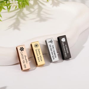 May include: Four rectangular medical alert tags in rose gold, gold, silver, and black finishes. Each tag features engraved medical symbols and personalized text including names, phone numbers, and health conditions like Type 1 Diabetes and allergies.