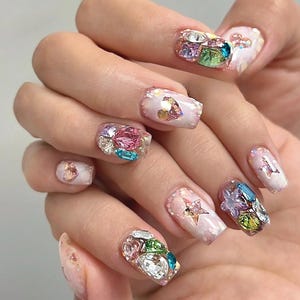 May include: Close-up of a hand with elaborately decorated nails. Each nail features a base of light pink polish with iridescent glitter and is adorned with colourful, sparkling rhinestones and gems. Some nails have star and heart accents.