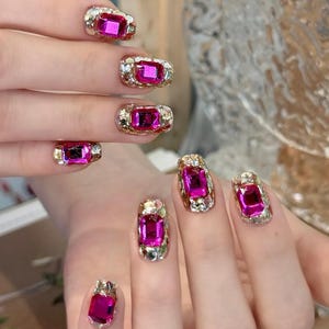 Może przedstawiać: Zbliżenie dłoni z manicure. Każdy paznokieć zdobi duży, prostokątny, różowy kamień szlachetny osadzony w złoto-srebrnym, inkrustowanym wzorze. Paznokcie to odważna i efektowna deklaracja modowa.