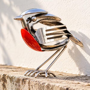 May include: A bird sculpture crafted from repurposed stainless steel cutlery, featuring a bright red painted spoon as the chest, a fork for the wing, and a small metal ball for the eye, standing on thin metal wire legs.