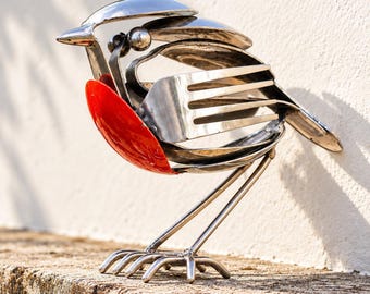 May include: A bird sculpture crafted from repurposed stainless steel cutlery, featuring a bright red painted spoon as the chest, a fork for the wing, and a small metal ball for the eye, standing on thin metal wire legs.