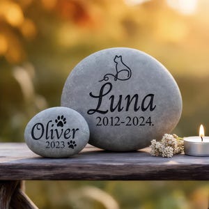 Peut inclure: Deux pierres tombales grises avec texte et graphiques noirs. L'une porte l'inscription "Luna 2012-2024" avec une silhouette de chat. L'autre porte l'inscription "Oliver 2023" avec des empreintes de pattes. Une bougie allumée et des fleurs sont à proximité.
