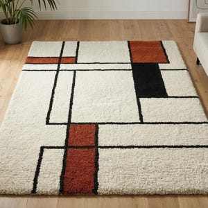 Puede incluir: Una alfombra rectangular con un diseño geométrico. La alfombra presenta un fondo crema con formas rectangulares negras, óxido y negras. La alfombra está colocada sobre un suelo de madera y un sofá es visible en el fondo.