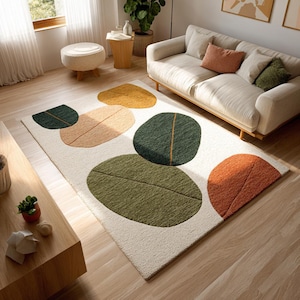 May include: A rectangular area rug with an off-white background and large, abstract leaf shapes in shades of green, orange, and yellow. The rug is placed on a hardwood floor, and a sofa and other furniture are visible in the background.