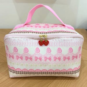 May include: A rectangular pink and white cosmetic bag with a top handle and a zipper closure. The fabric features a repeating pattern of strawberries, pink bows, and brown checkered trim. Two small red strawberry-shaped charms hang from the zipper.