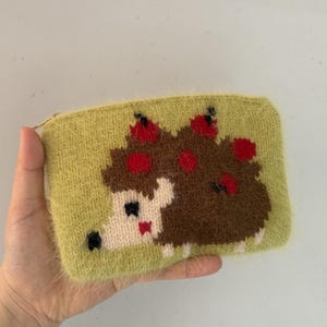 May include: A fuzzy, light green rectangular zippered pouch featuring a knitted graphic of a brown hedgehog with red apples on its back. The hedgehog has a cream-colored face, a black nose, and a small red mouth, all rendered in a soft, textured yarn.