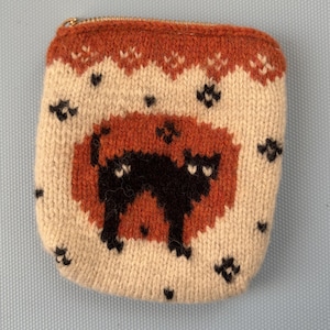 May include: A rectangular knit pouch featuring a cream-colored background with a central rust-orange oval containing a black cat silhouette. The design includes small black paw prints scattered across the cream area and a rust-colored border at the top.