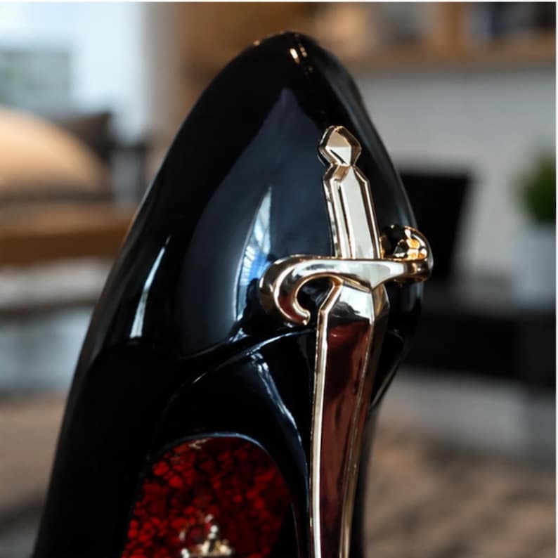 May include: A high-heeled shoe with a glossy black finish and a pointed toe. The heel is adorned with a metallic gold-toned sword ornament. The interior lining of the shoe features a textured red pattern visible near the heel base.