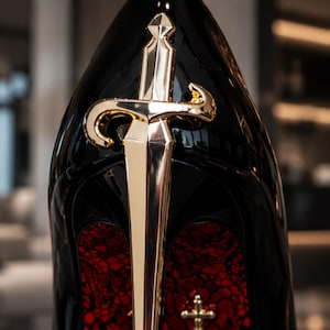 May include: A high-heeled shoe with a glossy black finish, featuring a prominent gold-toned sword emblem attached to the back of the heel. The interior lining of the shoe displays a textured red pattern with a small gold cross detail near the base.