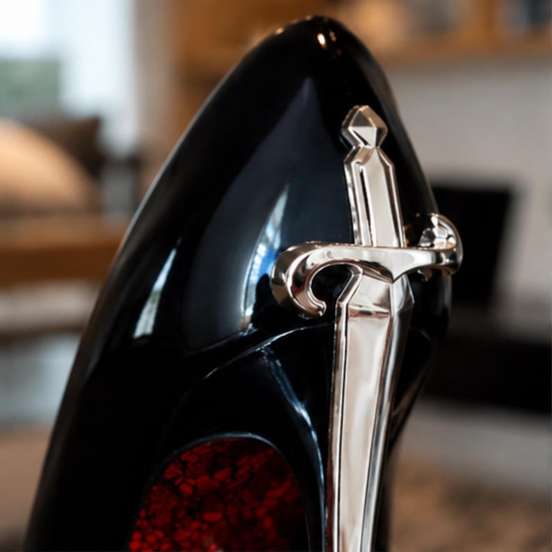 May include: A high-heeled shoe with a glossy black finish, featuring a prominent silver-toned sword emblem attached to the heel. The interior of the shoe displays a textured, deep red surface visible from the back angle of the footwear.