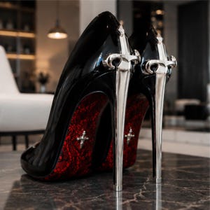 May include: A pair of black patent leather high-heeled shoes featuring silver-toned sword-shaped heels. The soles are detailed with a red lace pattern and a small silver cross emblem positioned on the arch of each shoe.