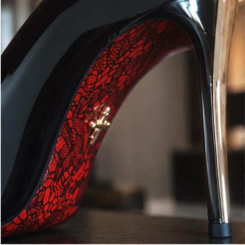 May include: A high-heeled shoe with a glossy black exterior and a metallic stiletto heel. The arch of the shoe features a vibrant red base layer covered by a black lace pattern, accented by a small, gold-toned cross-shaped emblem.