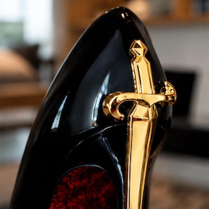 May include: A glossy black high-heeled shoe featuring a prominent, polished gold-tone sword emblem attached to the outer side. The interior of the shoe is lined with a textured, deep red material that contrasts with the smooth black exterior finish.