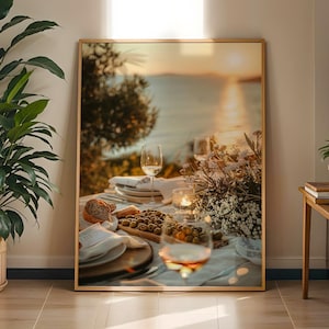 May include: A framed wall art print depicting an outdoor dining table set for a meal at sunset. The scene features a wooden board with green olives, a loaf of bread, white napkins, wine glasses, and a bouquet of white flowers near the sea.