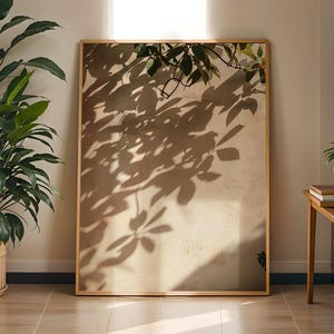 May include: A rectangular wall art piece in a thin gold frame featuring a photographic print of dark leaf shadows cast onto a textured beige wall. The image captures the contrast between the deep brown foliage silhouettes and the light surface.