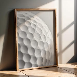 Può includere: Stampa incorniciata con un primo piano di una pallina da golf bianca con una superficie a fossette. La cornice è in legno chiaro. La stampa è appoggiata a una parete grigia e a un pavimento in legno.