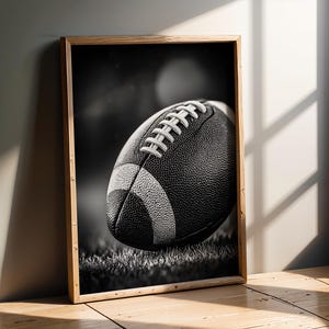 Può includere: Stampa incorniciata in bianco e nero di un pallone da football americano. Il pallone, con i suoi lacci bianchi e la superficie strutturata, è al centro. Lo sfondo è sfocato, suggerendo una partita. La cornice è in legno chiaro.