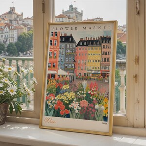 May include: A framed art print titled FLOWER MARKET with the text Wien and 2024. The illustration depicts a colorful European street scene with multi-story buildings in shades of orange, blue, and yellow, fronted by a vibrant garden of flowers.