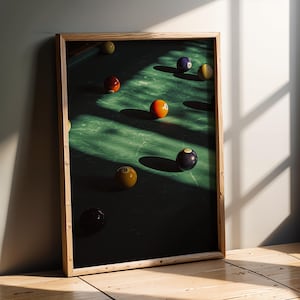 May include: A framed print featuring a close-up of a pool table with several billiard balls. The balls are various colors, including orange, yellow, and black, on a green felt surface. The wooden frame adds a classic touch to the artwork.